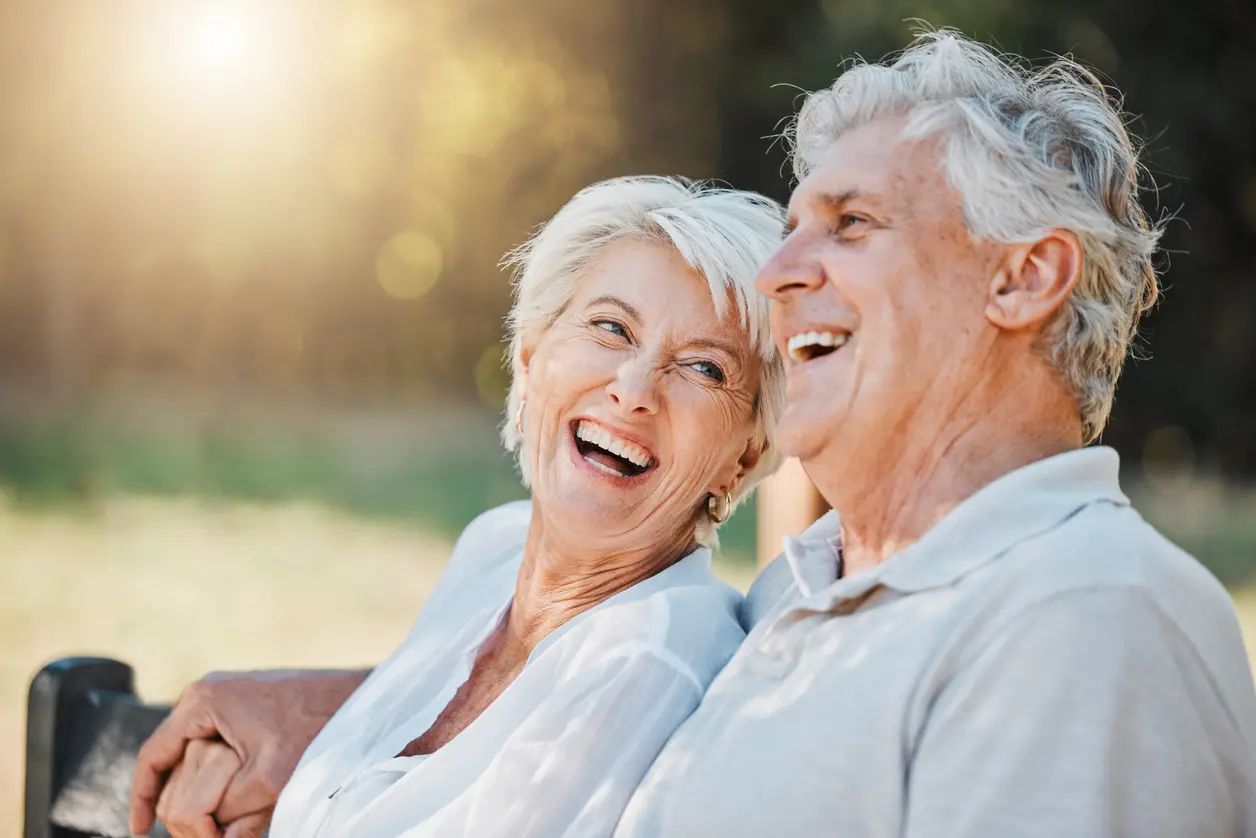 Ein älteres Paar sitzt auf einer Bank im Freien und lächelt sich an. Sie wirken glücklich und zufrieden, während sie miteinander lachen. Die Umgebung ist natürlich und sonnig, was eine freundliche und entspannte Atmosphäre schafft.