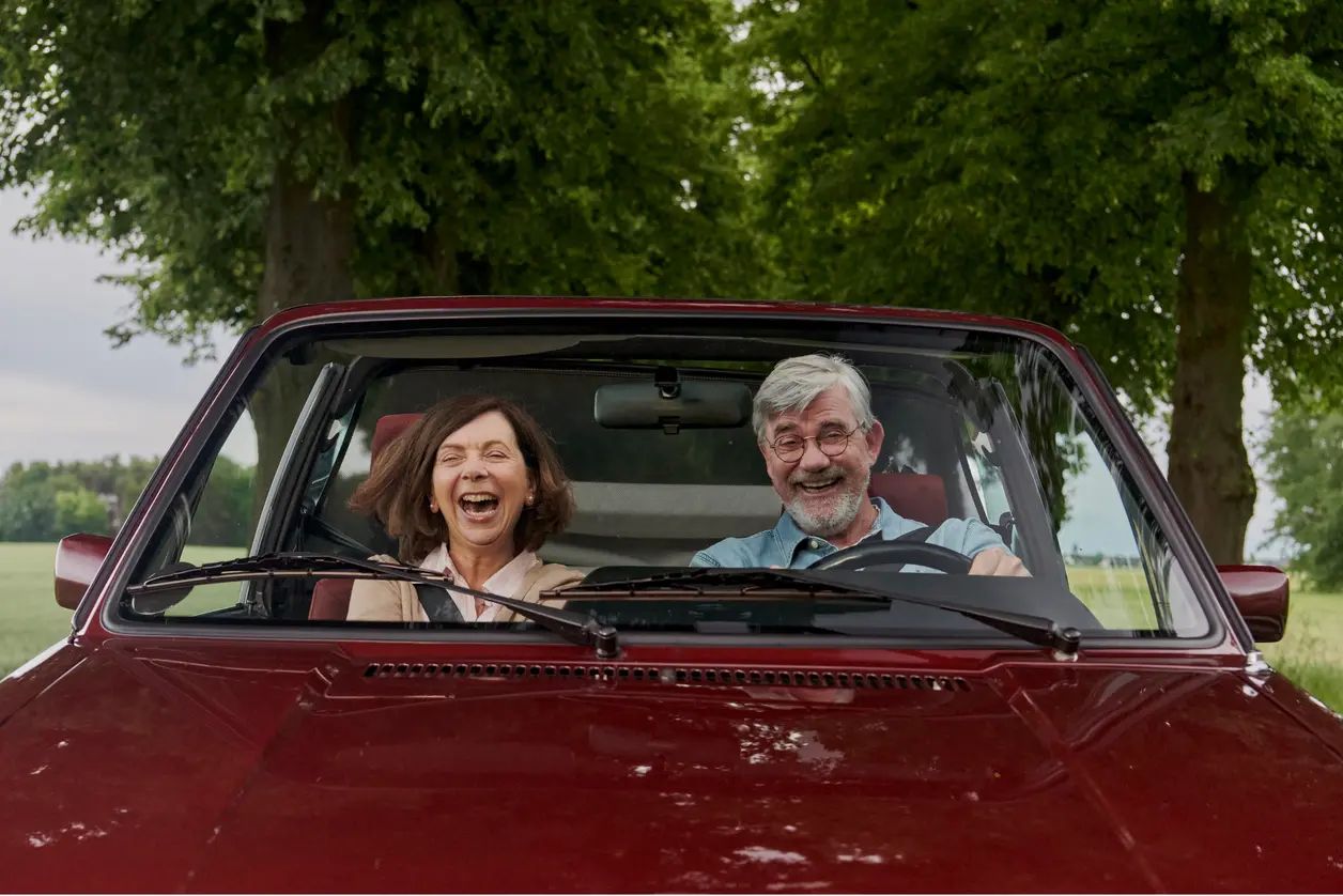 Älteres Paar fährt lachend in rotem Cabrio.
