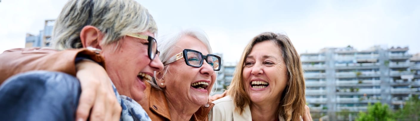 Drei Frauen mittleren Alters lachen gemeinsam, stehen eng beisammen und wirken fröhlich. Sie tragen Brillen und sind in einer städtischen Umgebung vor einem modernen Gebäudekomplex. Die Stimmung ist ausgelassen und freundlich.