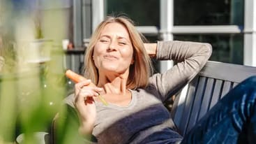 Eine Frau sitzt entspannt auf einer Bank im Freien, hält eine Karotte in der Hand und lächelt. Im Hintergrund sind unscharfe Blumen und ein modernes Gebäude zu sehen.