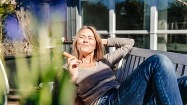 Eine Frau sitzt entspannt auf einer Bank im Freien, hält eine Karotte in der Hand und lächelt. Im Hintergrund sind unscharfe Blumen und ein modernes Gebäude zu sehen.