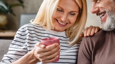 Ein lächelndes Paar sitzt auf einem Sofa. Die Frau mit blonden Haaren hält einen pinken Becher, während der Mann mit grauen Haaren sie anlächelt. Die Atmosphäre ist entspannt und freundlich, mit einem pflanzlichen Element im Hintergrund.