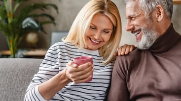 Ein lächelndes Paar sitzt auf einem Sofa. Die Frau mit blonden Haaren hält einen pinken Becher, während der Mann mit grauen Haaren sie anlächelt. Die Atmosphäre ist entspannt und freundlich, mit einem pflanzlichen Element im Hintergrund.
