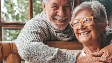 Ein älteres Paar sitzt nah beieinander auf einem Sofa, lächelt glücklich und umarmt sich. Der Mann hat graue Haare und trägt einen grauen Pullover, während die Frau graue Haare hat und eine Brille mit orangefarbenem Rahmen trägt.