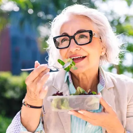 Eine ältere Frau mit grauen Haaren und Brille sitzt im Freien und lächelt, während sie mit einer Gabel einen Salat aus einem Behälter isst. Die Umgebung ist grün und sonnig.