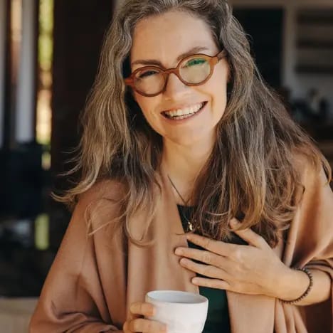 Eine lächelnde Frau mit langen, lockigen Haaren und Brille trägt einen beigen Überwurf. Sie hält eine Tasse in der Hand und steht in einem hellen Raum mit einem warmen, einladenden Ambiente.