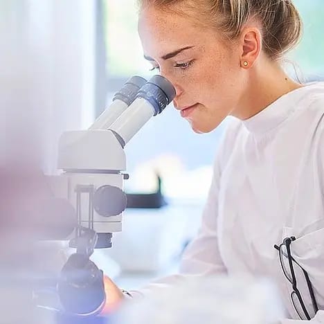 Eine Wissenschaftlerin betrachtet durch ein Mikroskop, während sie an einem Tisch arbeitet. Sie trägt eine weiße Laborkleidung und hat die Haare zurückgebunden. Der Hintergrund ist unscharf, was den Fokus auf die Mikroskopnutzung lenkt.