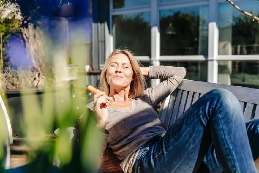 Eine Frau sitzt entspannt auf einer Bank im Freien, hält eine Karotte in der Hand und lächelt. Im Hintergrund sind unscharfe Blumen und ein modernes Gebäude zu sehen.