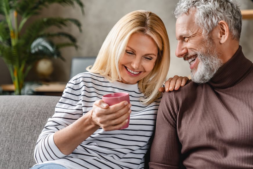 Ein lächelndes Paar sitzt auf einem Sofa. Die Frau mit blonden Haaren hält einen pinken Becher, während der Mann mit grauen Haaren sie anlächelt. Die Atmosphäre ist entspannt und freundlich, mit einem pflanzlichen Element im Hintergrund.